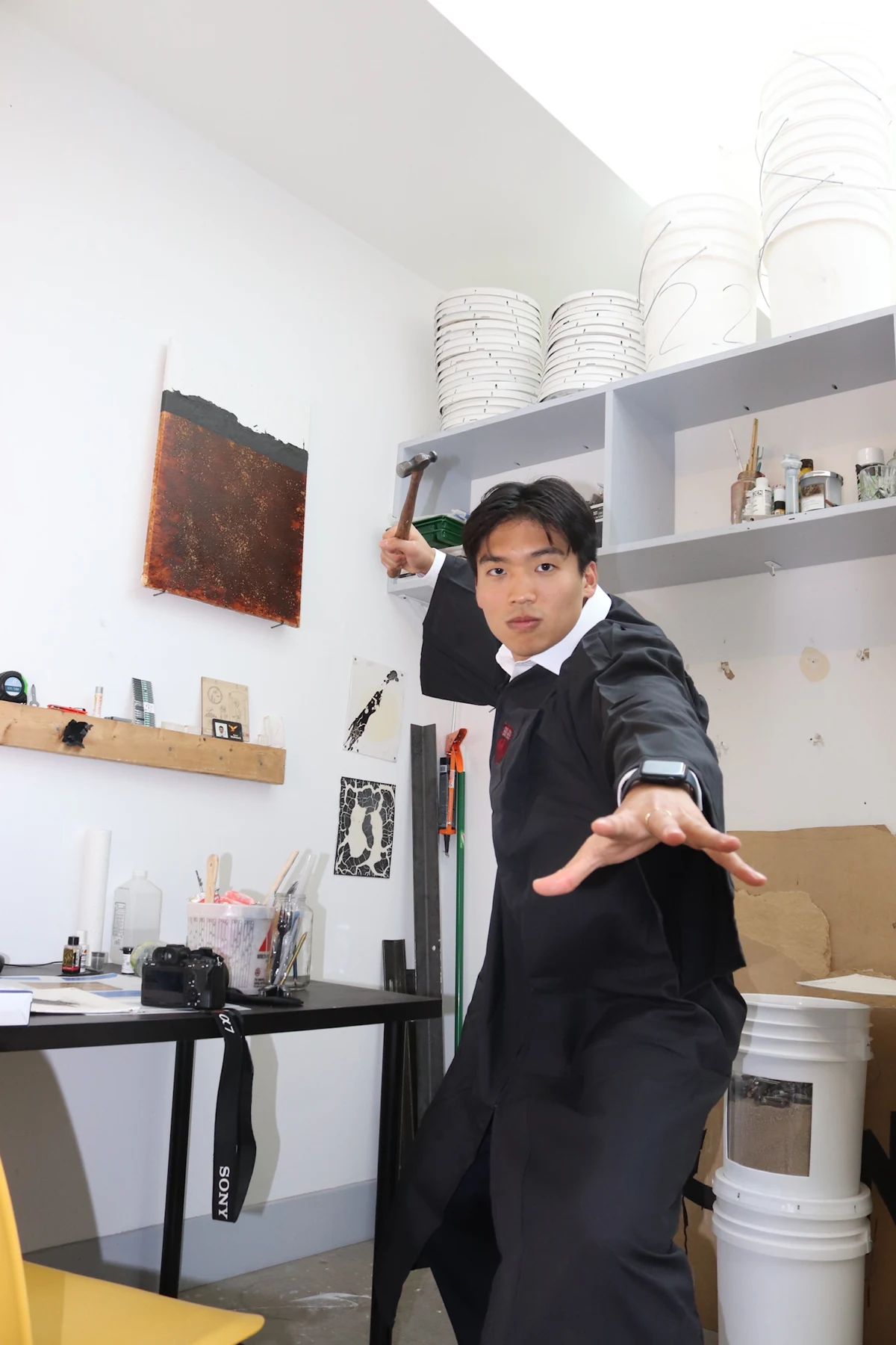 Thom striking a playful pose with a hammer in graduation robes in the studio